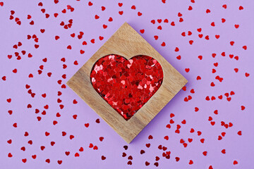 Wooden heart-shaped box with red sequins in form of hearts on purple background with scattered random heart-shaped sequins