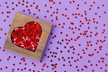Wooden heart-shaped box with red sequins in form of hearts on purple background with scattered random heart-shaped sequins
