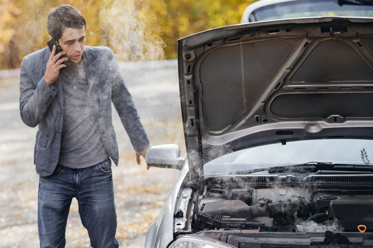 A man looks under the open hood of a car. The car broke down on the road. The engine is smoking