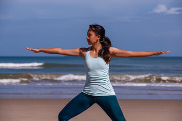 people, fitness, sport and healthy lifestyle concept - young asian woman making yoga warrior pose on tropical beach and blue sky background