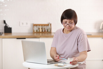 Concentrated senior woman in eyeglasses calculating utility bills or domestic expenditures,doing financial paperwork,paying for services, insurance using computer e-banking application alone at home.