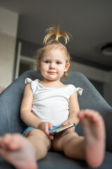 Smiling little girl is sitting on armchair and using smart phone