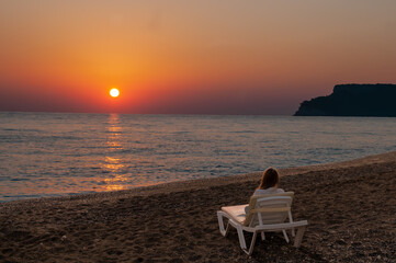 Woman sitting on a sun lounger on the beach against the backdrop of a sea sunset