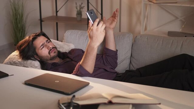 Mobile communication. Relaxed man. Study break. Handsome guy laying sofa having video call on smartphone educational goods on desk light shadow room interior.