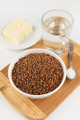 Raw buckwheat in a plate, a glass of water, oil and salt on a cutting board. Ingredients for cooking. Healthy, simple food.
