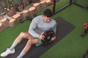 A fit asian man doing weighted Russian twists with a barbell plate while sitting on a black mat. Core workout at his home gym.