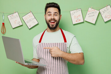 Young smiling fun male chef confectioner baker man 20s in striped apron hold use work point on laptop pc computer isolated on plain pastel light green background studio portrait. Cooking food concept.