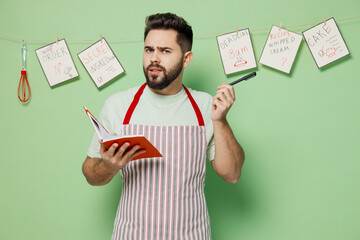 Young confused puzzled male chef confectioner baker man in striped apron write down notes recipe...