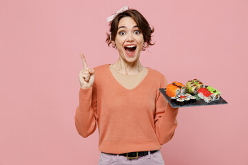 Young fun woman 20s in casual clothes hold in hand makizushi sushi roll served on black plate traditional japanese food holding index finger up great new idea isolated on plain pastel pink background © ViDi Studio