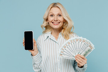 Elderly smiling fun woman 50s wear striped shirt holding fan of cash money in dollar banknotes use mobile cell phone with blank screen workspace area isolated on plain pastel light blue background