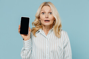 Elderly shocked surprised woman 50s wearing striped shirt holding in hand use mobile cell phone with blank screen workspace area isolated on plain pastel light blue color background studio portrait