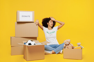 Full body tired young woman of African American ethnicity wear white volunteer t-shirt sit near boxes with presents isolated on plain yellow background. Voluntary free work assistance help concept