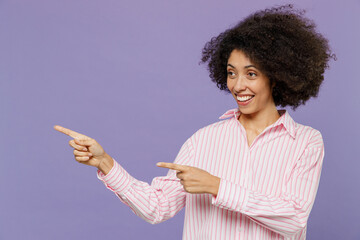 Young cool woman of African American ethnicity 20s in pink striped shirt point index finger aside on workspace area mock up copy space isolated on plain pastel light purple background studio portrait.