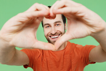 Close up young man 20s in casual orange t-shirt showing shape heart look through hands heart-shape sign isolated on plain pastel light green color background studio portrait. People lifestyle concept.