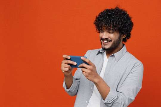 Gambling Jubilant Young Bearded Indian Man 20s Wear Blue Shirt Using Play Racing App On Mobile Cell Phone Hold Gadget Smartphone For Pc Video Games Isolated On Plain Orange Background Studio Portrait