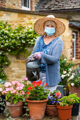Woman watering plants and flowers in the garden wearing a face mask covering during Coronavirus Covid-19 pandemic in lockdown