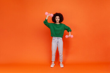 Full length portrait of woman with Afro hairstyle wearing green casual style sweater training biceps, using dumbbells, doing exercises for hands. Indoor studio shot isolated on orange background.