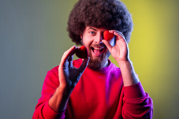 Hipster man with Afro hairstyle having fun with red toy hearts, showing his fondness and devotion, romance, wearing red sweatshirt. Indoor studio shot isolated on colorful neon light background.