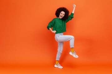 Fototapeta na wymiar Full length portrait of woman with Afro hairstyle wearing green casual style sweater screaming with happy expression, celebrating success. Indoor studio shot isolated on orange background.