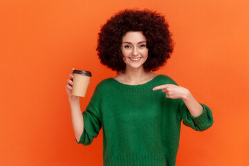Portrait of smiling woman with Afro hairstyle wearing green casual style sweater standing pointing at disposable cup with take away coffee. Indoor studio shot isolated on orange background.