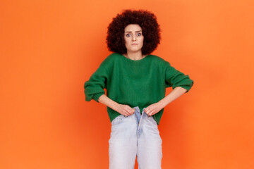 Portrait of upset sad young adult woman with Afro hairstyle wearing green casual style sweater unable to button jeans due to weight gain. Indoor studio shot isolated on orange background.