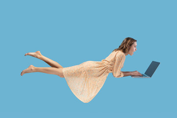Hovering in air. Surprised excited girl yellow dress levitating with laptop, typing keyboard, reading shocking news on laptop screen while flying in mid-air. studio shot isolated on blue background - obrazy, fototapety, plakaty