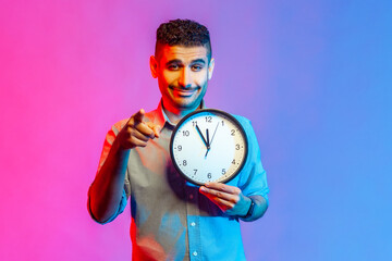 Portrait of smiling man in shirt pointing finger at camera holding big wall clock in hand, time to...