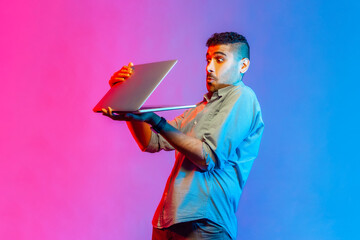 Portrait of young adult man in shirt holding laptop in hands, sees shocking scared content, looking...