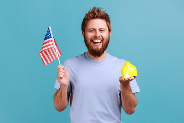 Portrait of happy handsome bearded man holding flag of united states of america and paper house, dreaming about living in usa, green card. Indoor studio shot isolated on blue background.