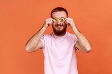 Fototapeta premium Portrait of smiling happy bearded man covering eyes with golden bitcoin, looking at camera through btc coin, wearing pink T-shirt. Indoor studio shot isolated on orange background.