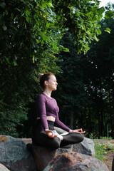 Mental health, harmony, mindfulness practice outdoors. Smiling cute young woman with closed eyes practicing meditation, caucasian yogi woman relaxing sitting in lotus position on stone in park