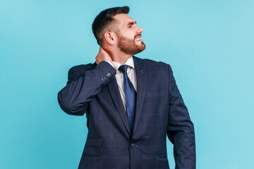 Portrait of worried anxious bearded man wearing official style suit touching neck, feeling acute pain moving and turning head, suffering spine problems. Indoor studio shot isolated on blue background.