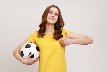 Young beautiful brown haired teenager girl holding soccer ball, looking at camera with toothy...