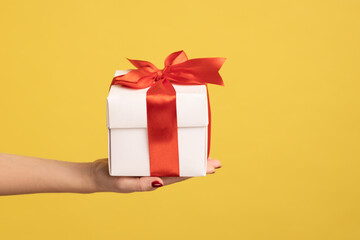 Closeup side view of woman hand holding out white gift box with red ribbon, giving present on...