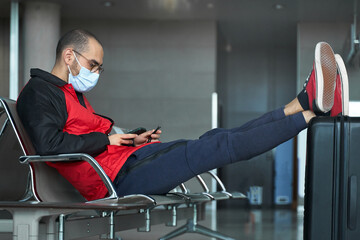 Young man with face mask using smartphone while waiting at airport with luggage during covid...