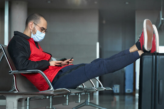 Young man with face mask using smartphone while waiting at airport with luggage during covid pandemic