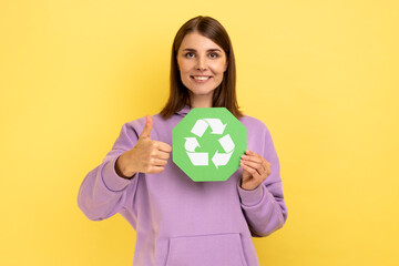 Positive woman with dark hair holding in hands green recycling sing, ecology concept, looking at camera and showing thumb up, wearing purple hoodie. Indoor studio shot isolated on yellow background.