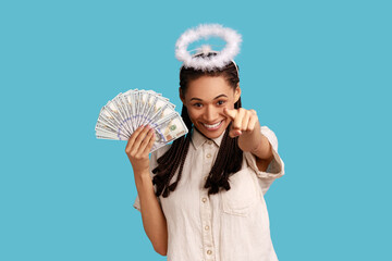 Positive angelic woman with black dreadlocks and halo over head holding fan of dollar bills and pointing finger to camera, choosing lottery winner. Indoor studio shot isolated on blue background.