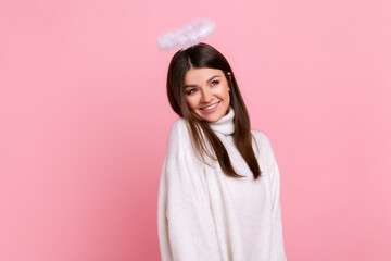 Attractive brunette angelic woman with nimb over head, looking away with dreamy facial expression, wearing white casual style sweater. Indoor studio shot isolated on pink background.