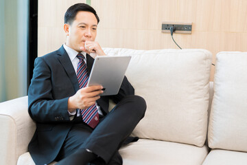 Asian businessman, wearing a suit and good looking, is checking information on a tablet.  On the sofa in the office at the company