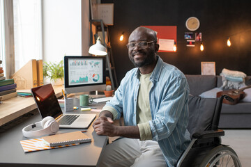Portrait of African man with disability sitting on wheelchair and smiling at camera while working...