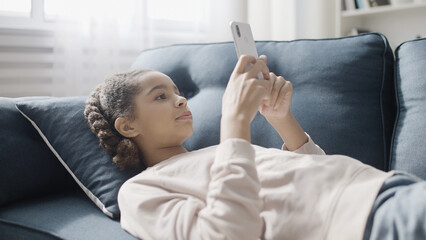 Cute black girl using social network app in smartphone, lying on sofa at home