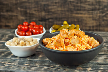 Pilaf with meat and carrots in a black bowl. Marinated mushrooms, tomato and cucumber on a black background. Food on a shabby table. Bowl on an old wooden board.