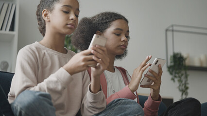 Two teenage girls sitting on couch with smartphones, browsing internet at home