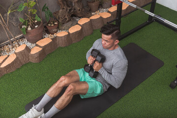 A fit asian guy sitting down on a black mat with two hex dumbbells. Starting position for dumbbell...