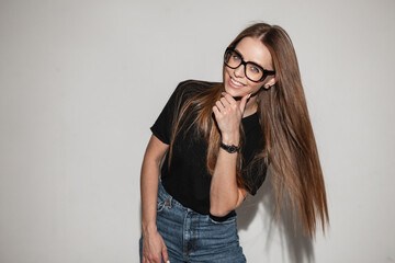 Beautiful happy young woman with stylish glasses in a trendy black t-shirt near a white wall....
