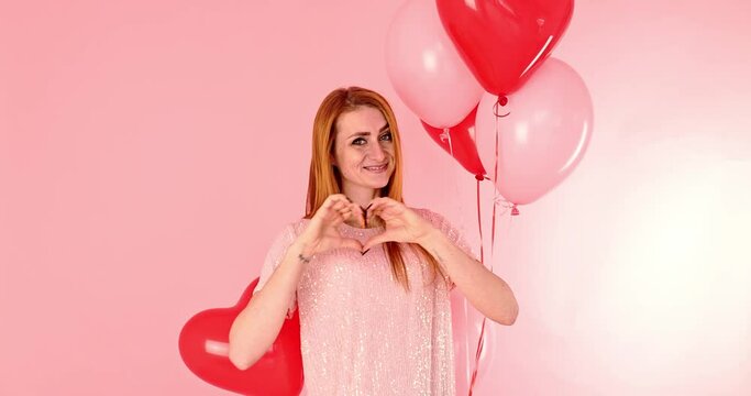 Beautiful redhead girl forming a heart with her hands with red heart balloon posing. Happy Valentine's Day concept. Studio photo of beautiful ginger girl dancing on pink background.