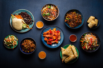 Set of assorted Chinese dishes on table: sweet and sour chicken on plate, fried spring rolls, noodles, salad, rice, steamed buns with bbq glazed pork, Asian style banquet or buffet, top view 
