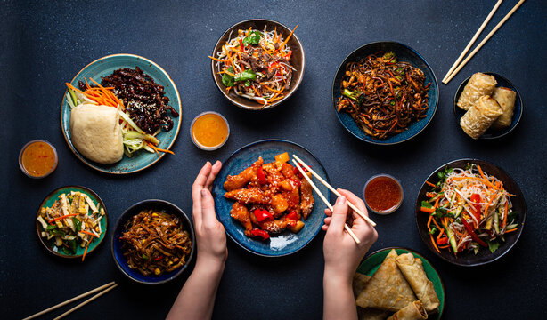 Set of Chinese dishes on table, female hands holding chopsticks: sweet and sour chicken, fried spring rolls, noodles, rice, steamed buns with bbq glazed pork, Asian style banquet or buffet, top view 
