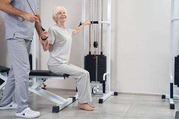 Fototapeta premium Pleased elderly woman pulling handle and cable of strength machines